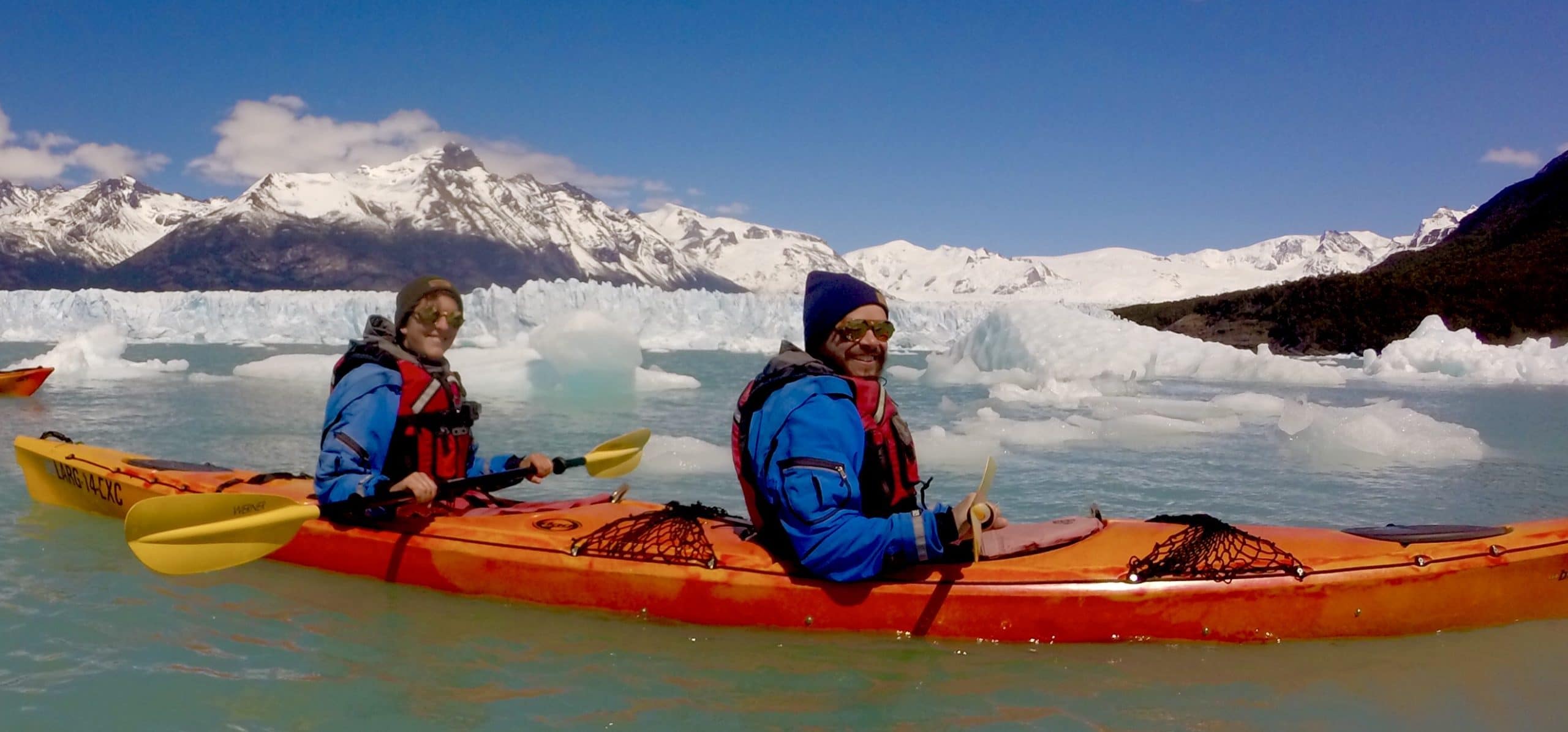 Myriam on the back and Pierre on the front of an orange kayak. They are dressed warmly and the water is icy with snowy mountains in the background.