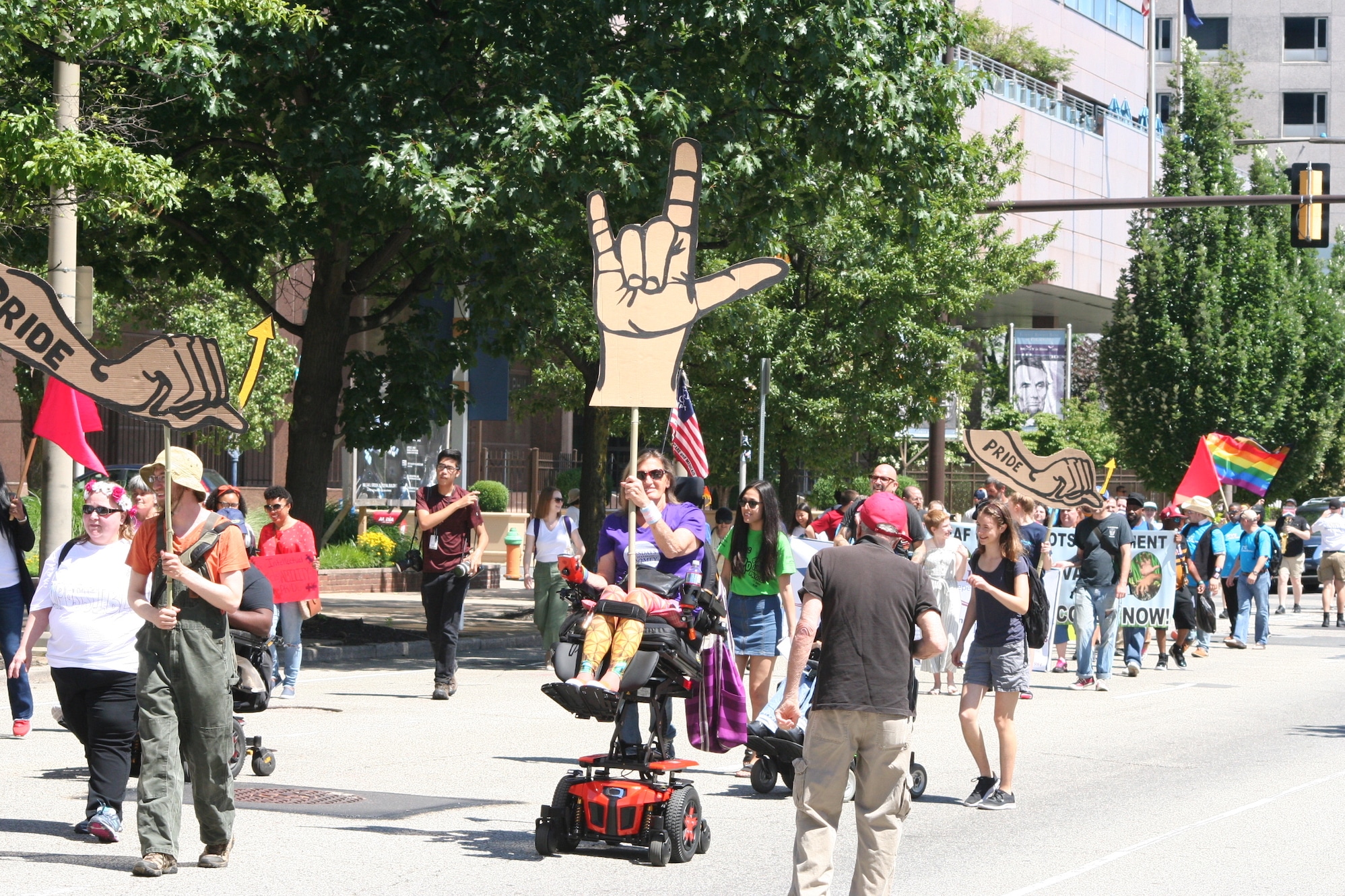 A woman in a power wheelchair holds a sign showing "I love you" in American Sign Language. Many people are marching and rolling behind her holding various signs.