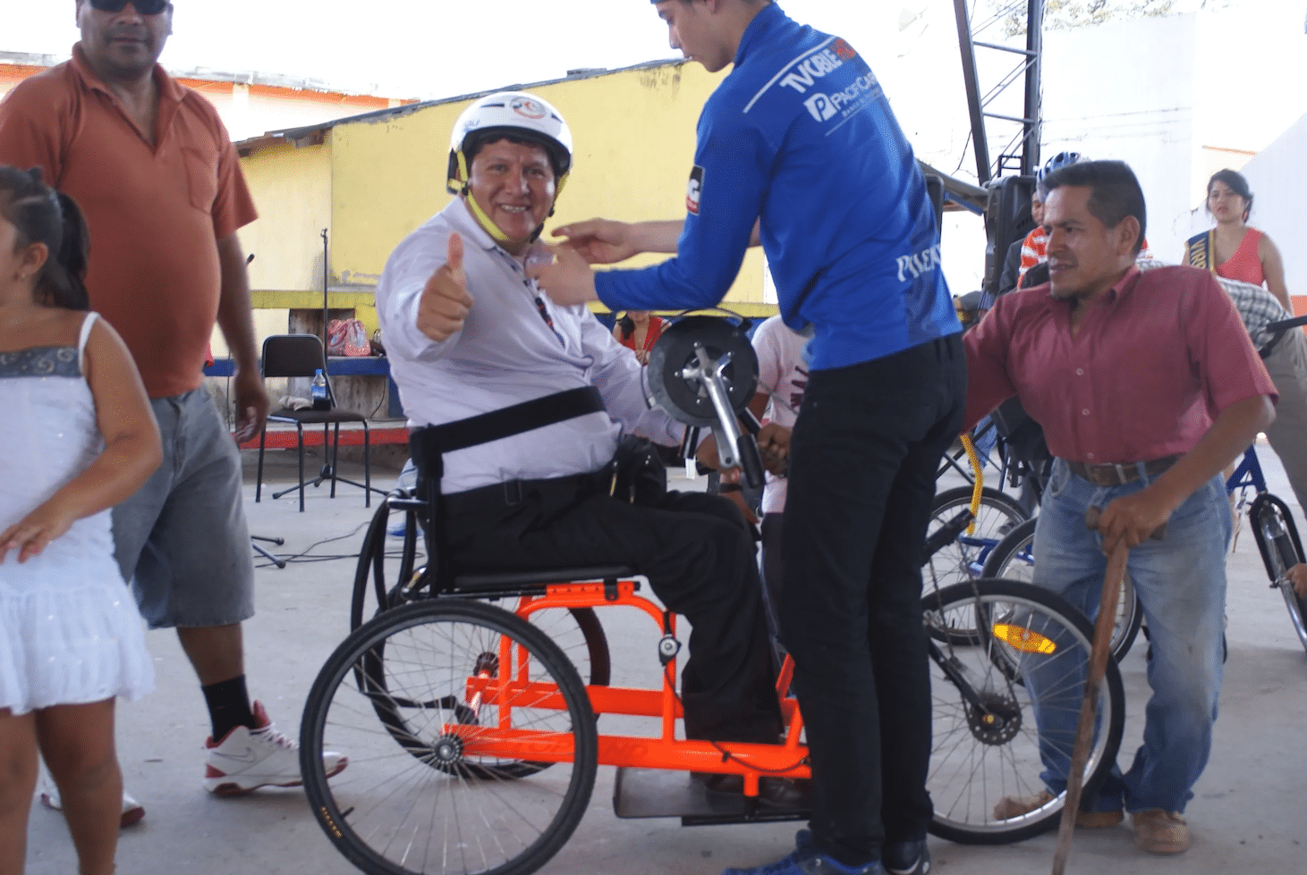 A man sits on an adaptive bike while an assistant straps him in. He's looking at the camera holding his thumb up.