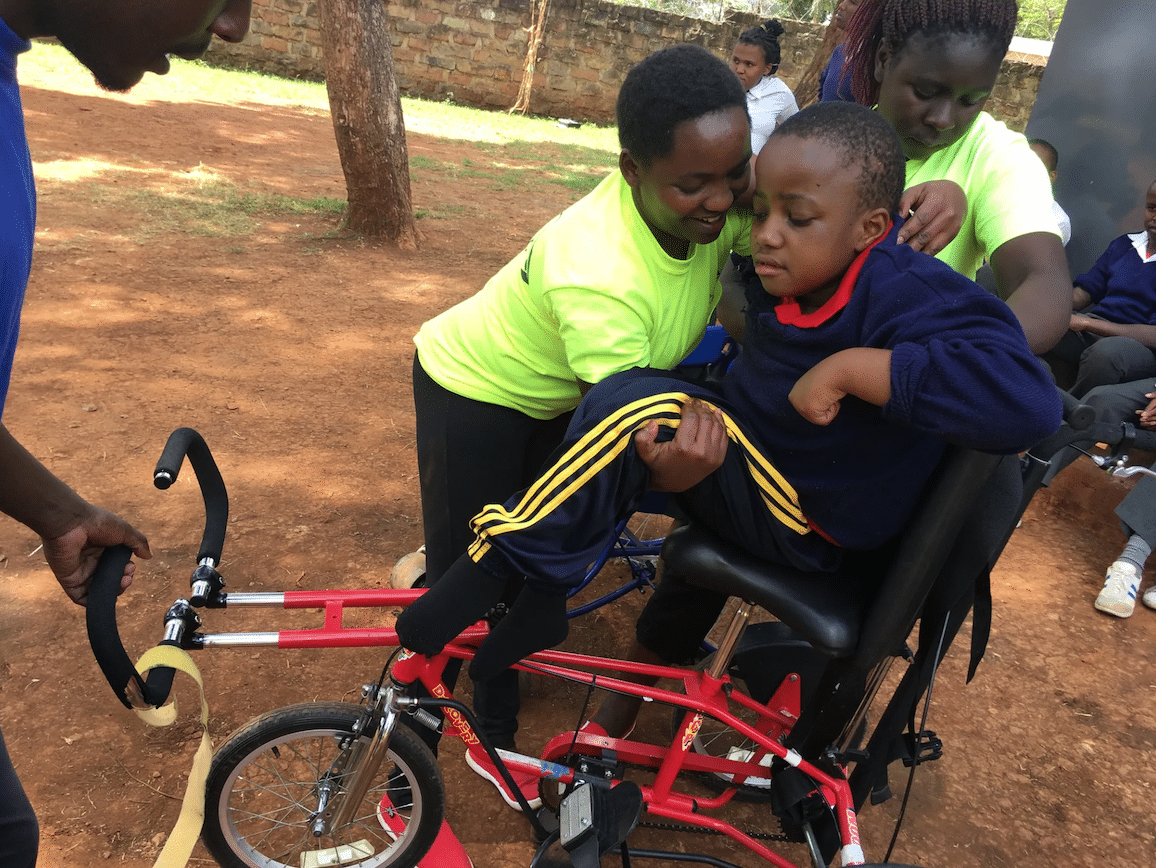 A young African boy lifts another young African boy onto a red adaptive bike.