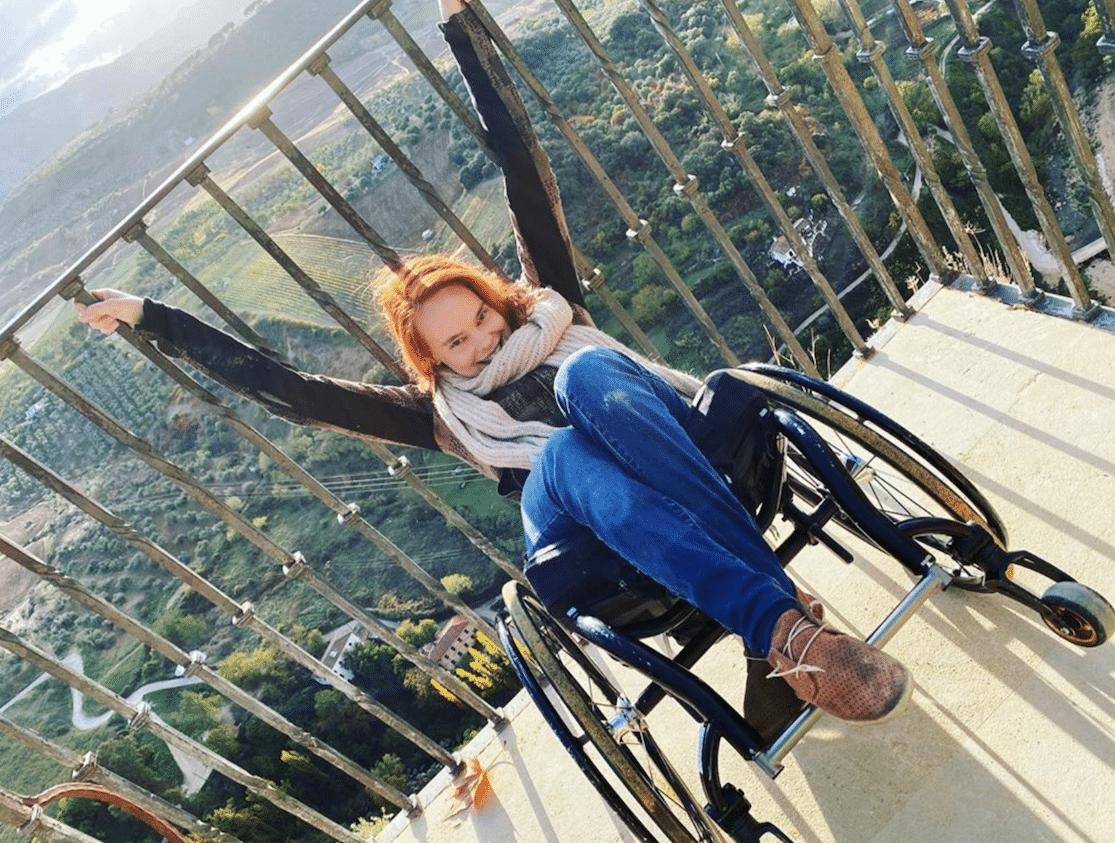 Erin Clark does a wheelie in her wheelchair while hanging onto a railing that overlooks a luscious, green landscape. She is smiling at the camera. 