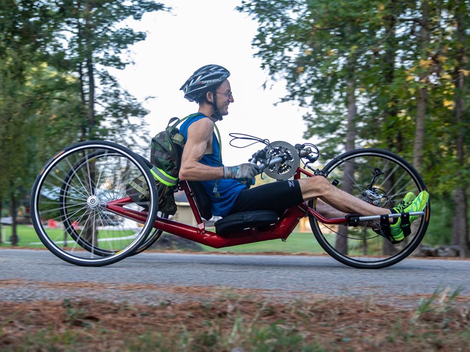 A side view of Jayden smiling while riding his handcycle on a cement path outside with trees all around..