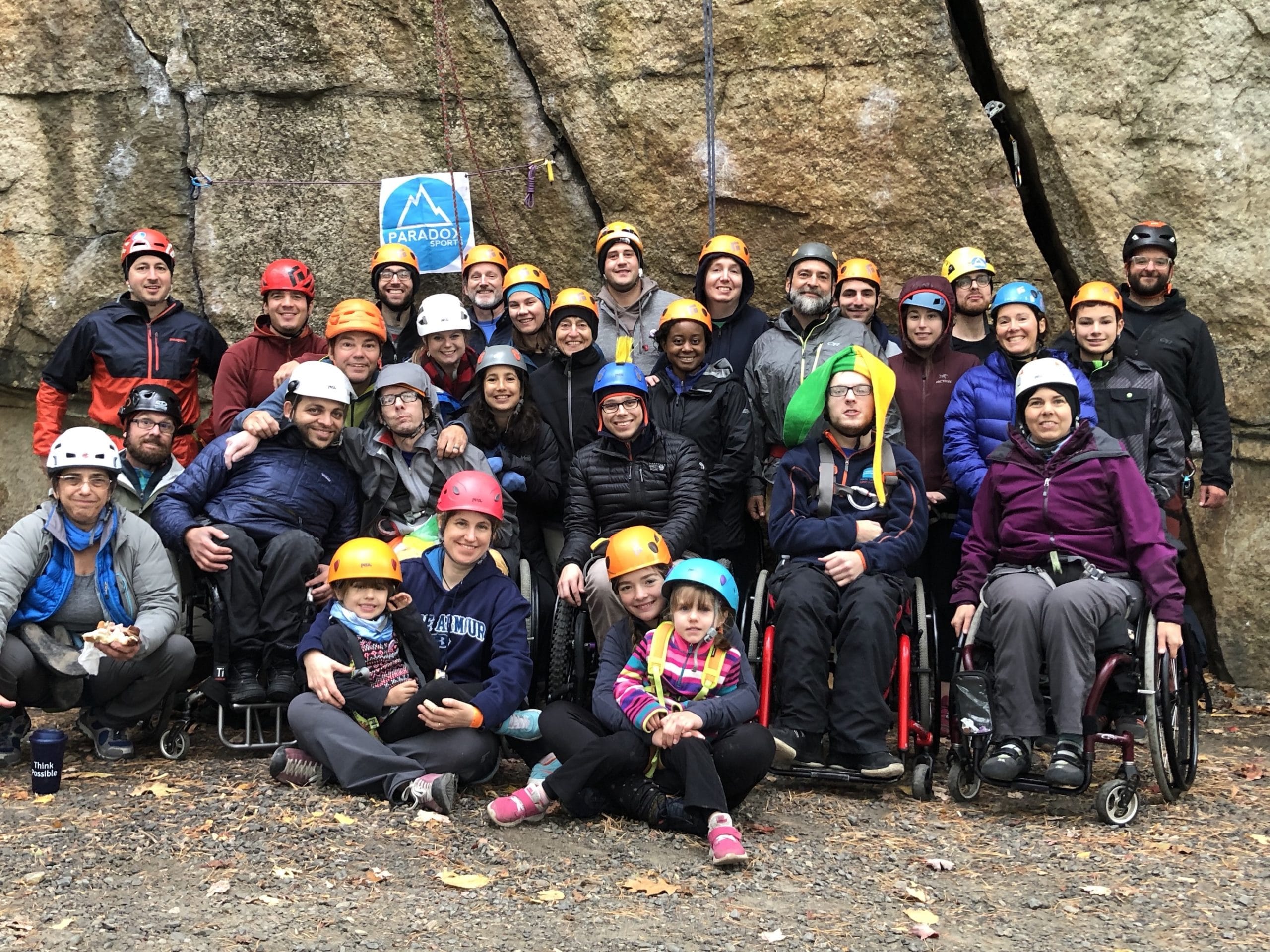 A group of climbers with and without disabilities wearing safety helmets in front of a cliff smiling at the camera.