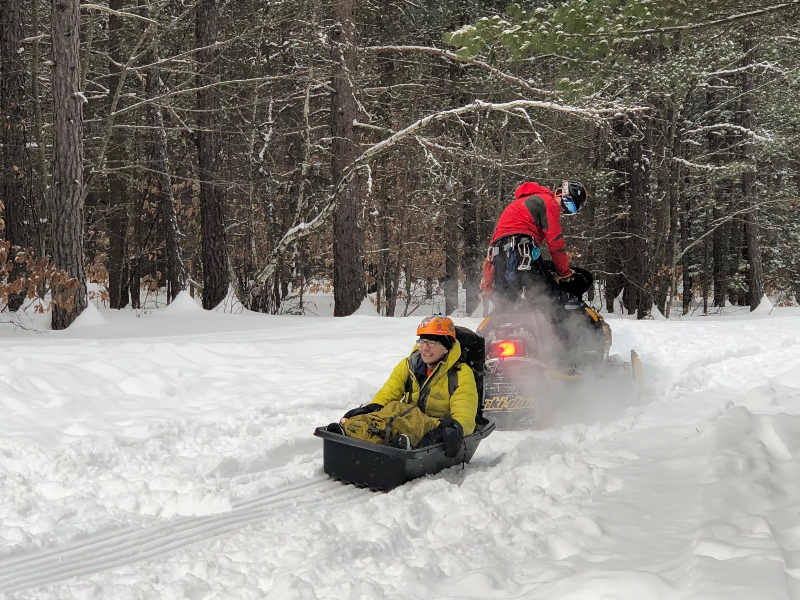 A snowmobile pulls a sled holding a man in a yellow snowsuit and orange helmet. 