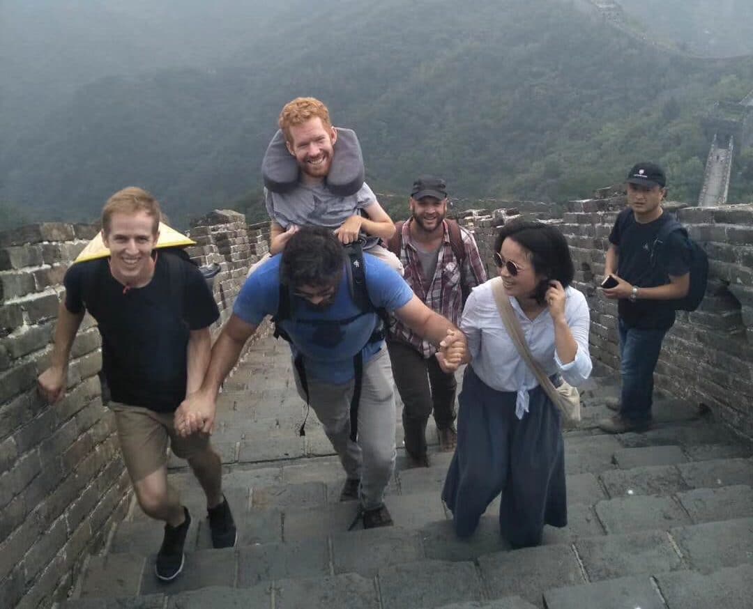 A man carries Kevan in a backpack up some stairs on the Great Wall of China. A man and a woman lend their hands to support the man carrying Kevan as two other men look on.