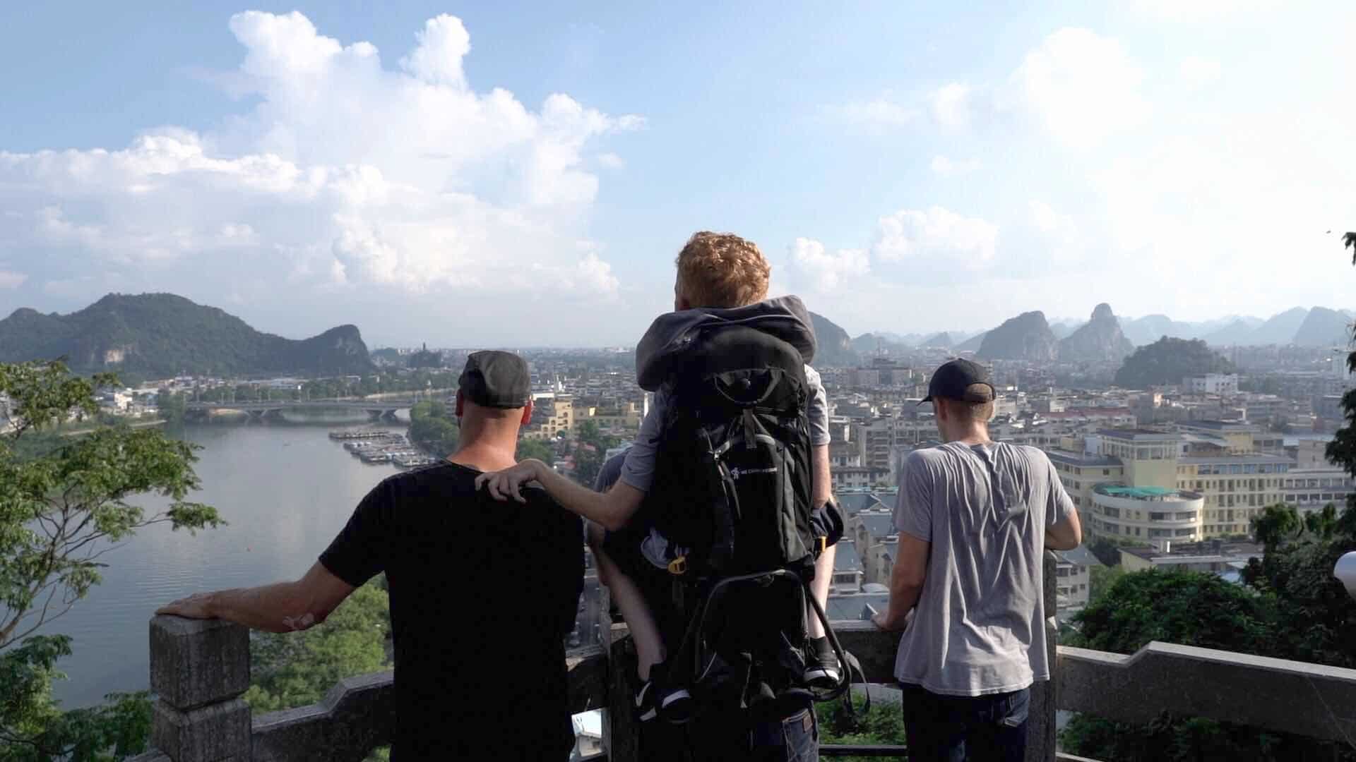 Four young men looking out over a European city on a river. One of the men is in a backpack on the back of the man in the middle. 