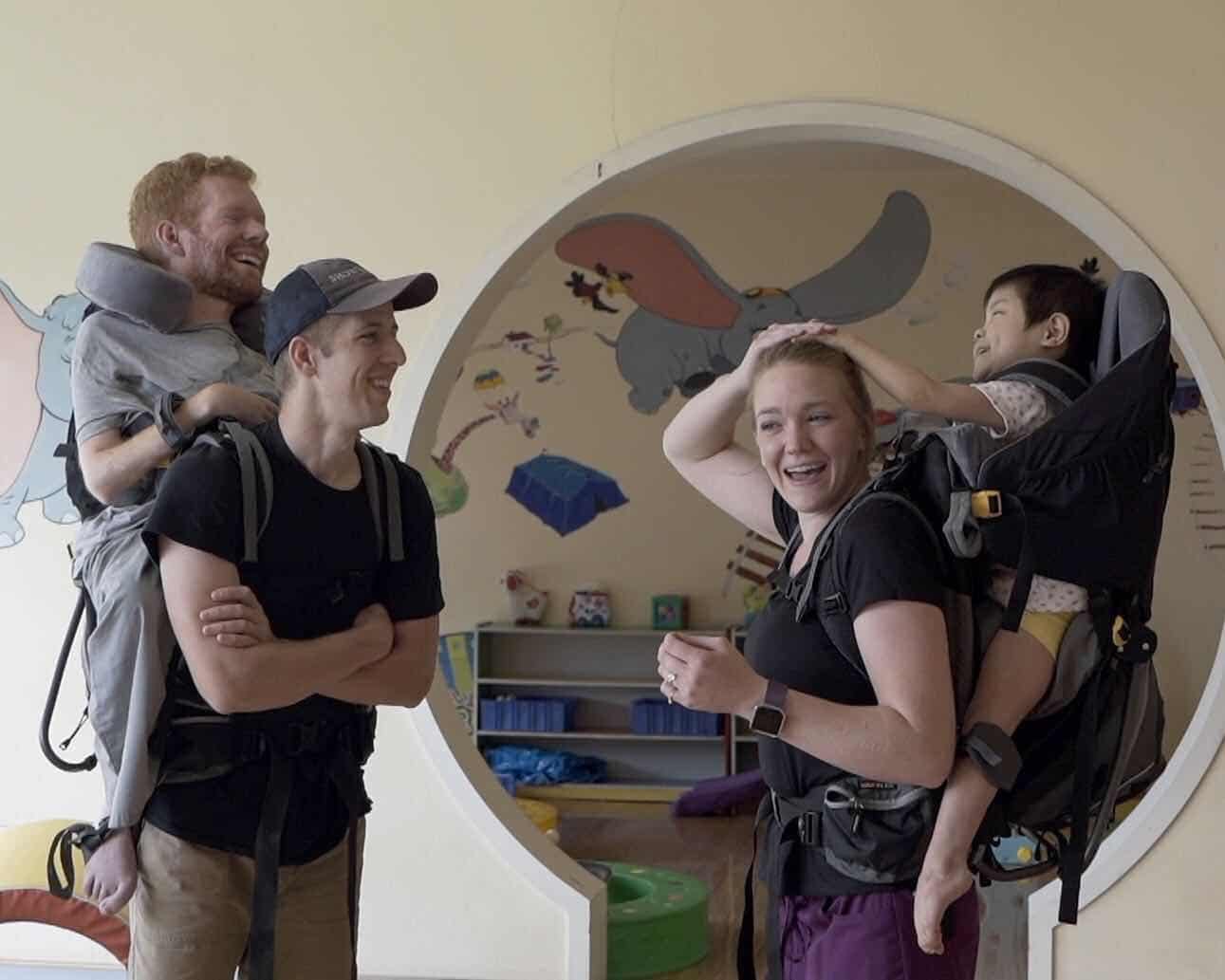 A man carrying Kevan in his backpack. Across from him stands a woman carrying a child in a backpack. The child has its hand on top of the woman's head and they're both laughing.