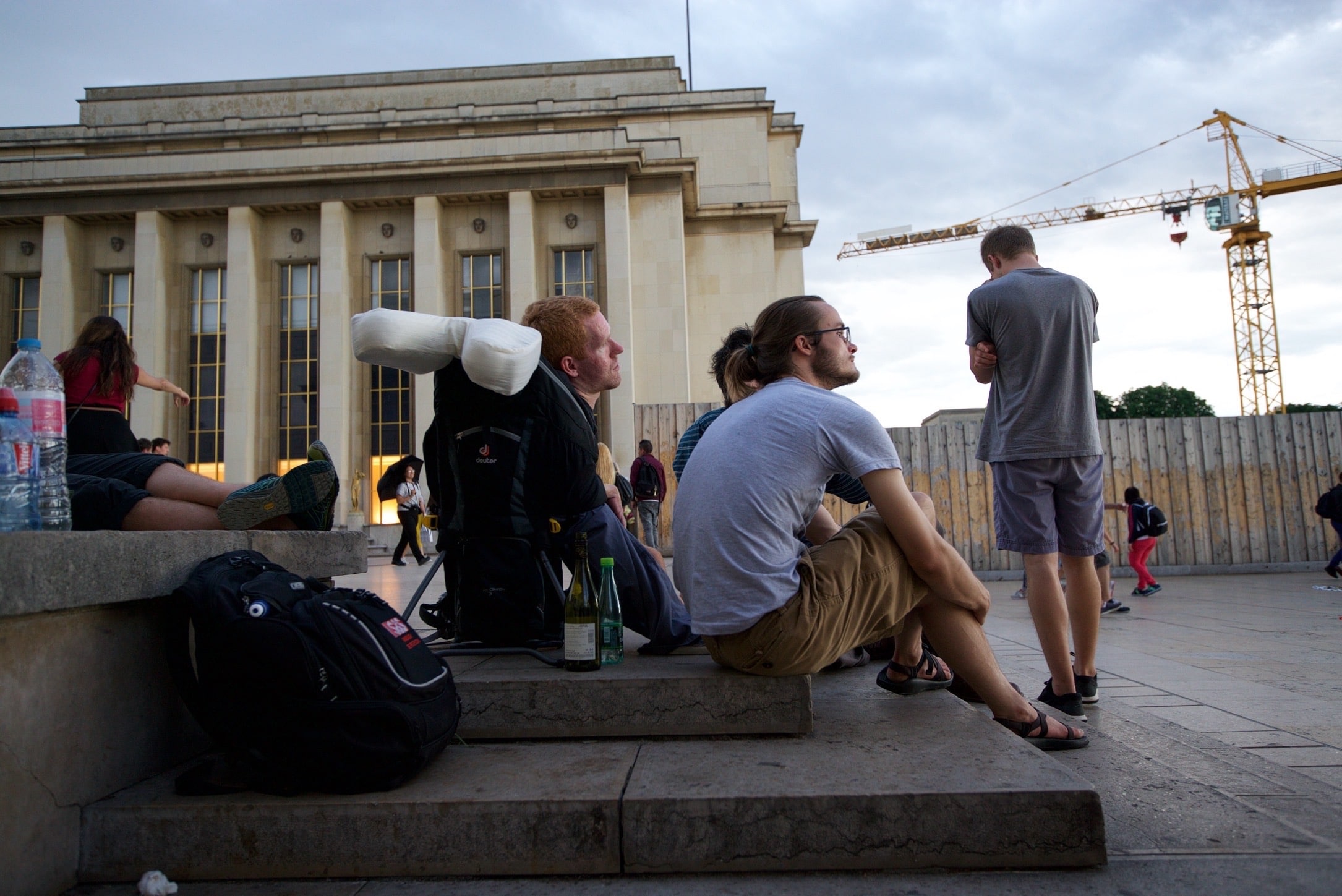 Kevan Chandler sitting in his carrying backpack on a set of cement steps in front of a large building with pillars. Other people are sitting on the steps and walking through the area.