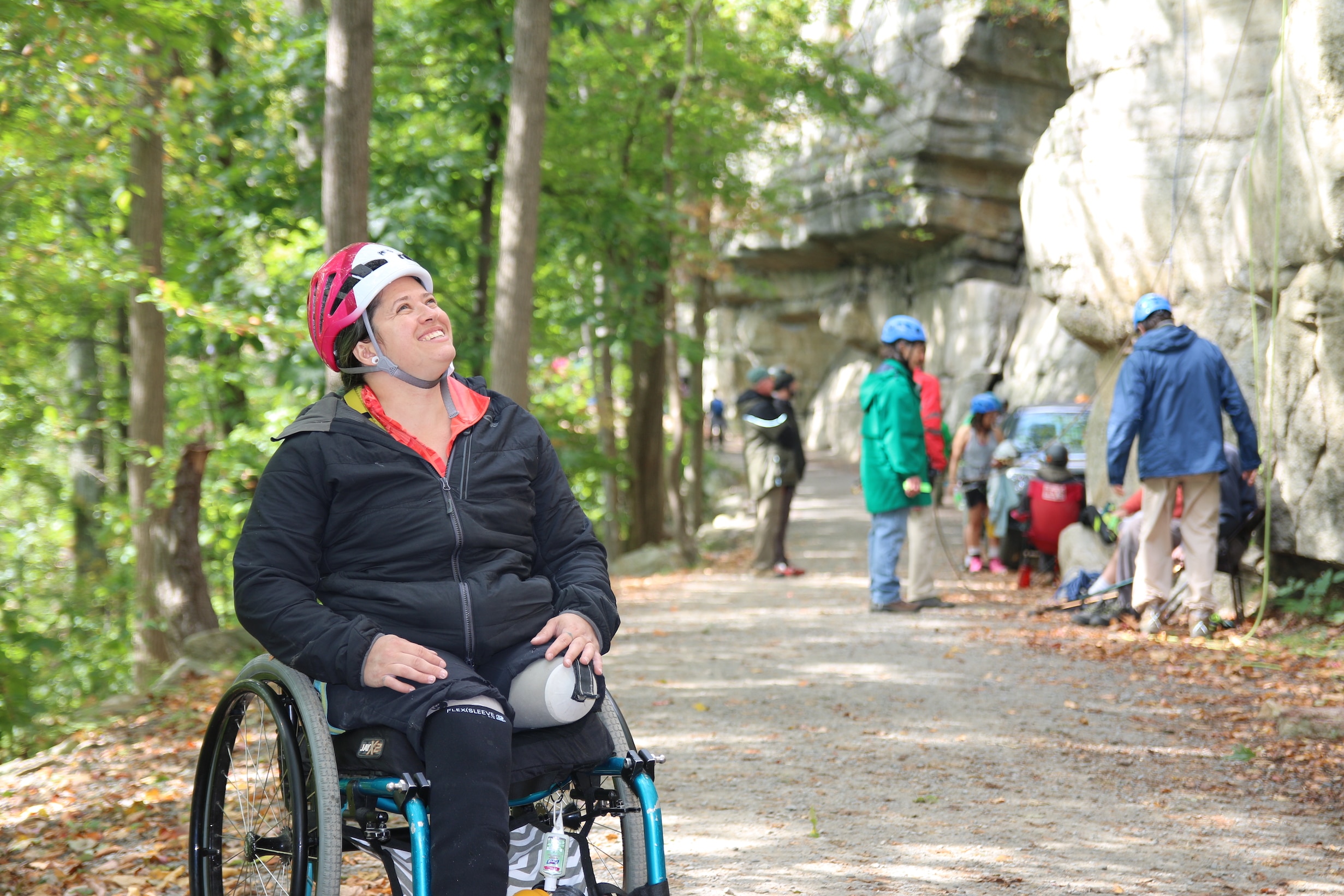 A woman single-leg amputee in a wheelchair wearing a helmet looks up toward the mountain and smiles. In the background, people prepare their climbing equipment.