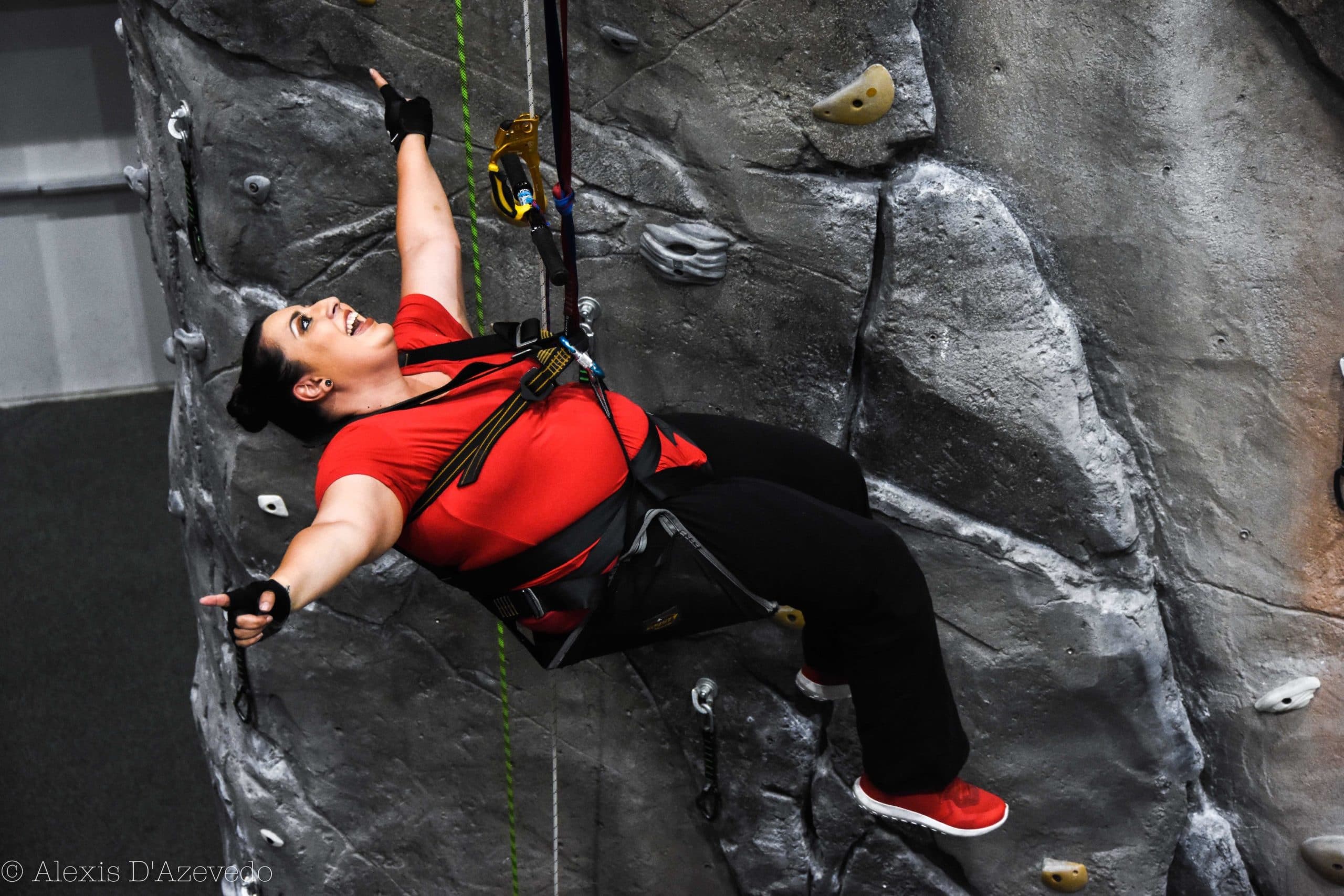 A woman outstretches her arms and leans back in her climbing chair as if to celebrate making it up the indoor wall.