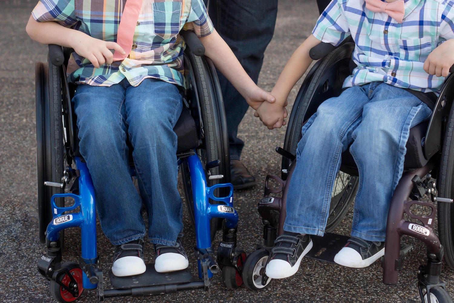 Two boys from the chest down in wheelchairs holding hands.