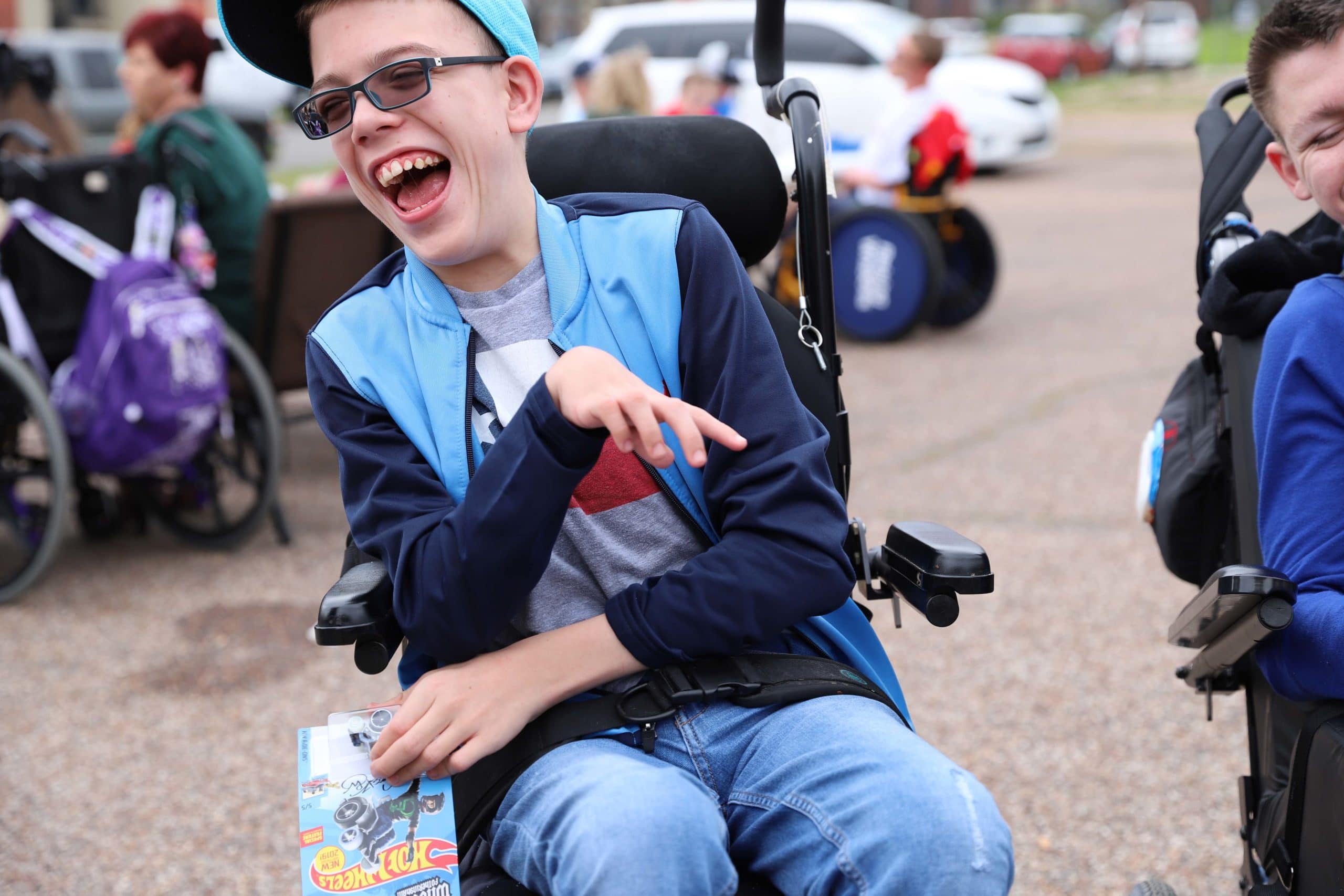 A boy in a power wheelchair wearing a baseball cap and sunglasses smiles large while looking off to his side.