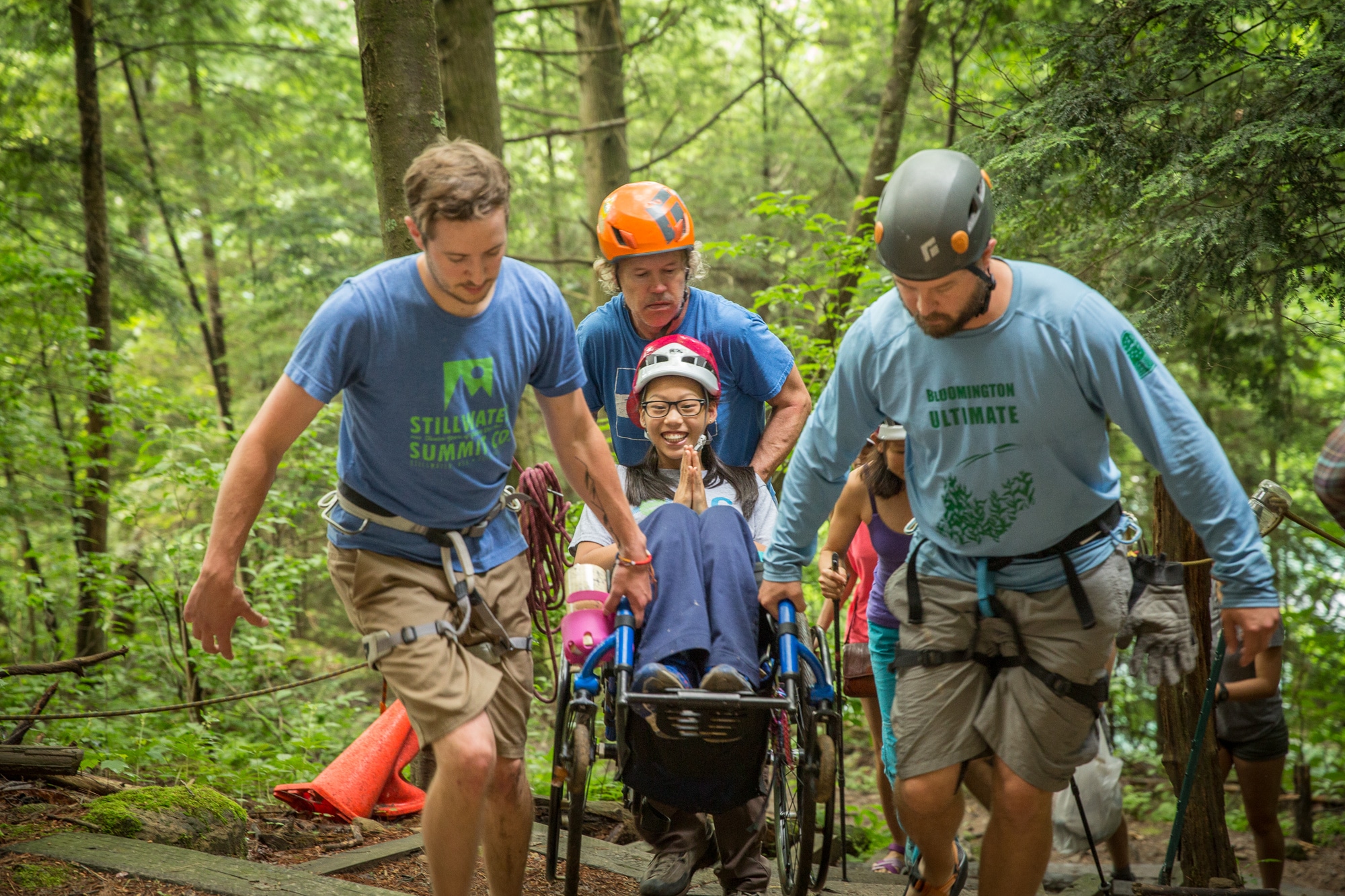 Three men wearing climbing equipment carry a smiling girl in her wheelchair up a set of wooden stairs in the forest. 